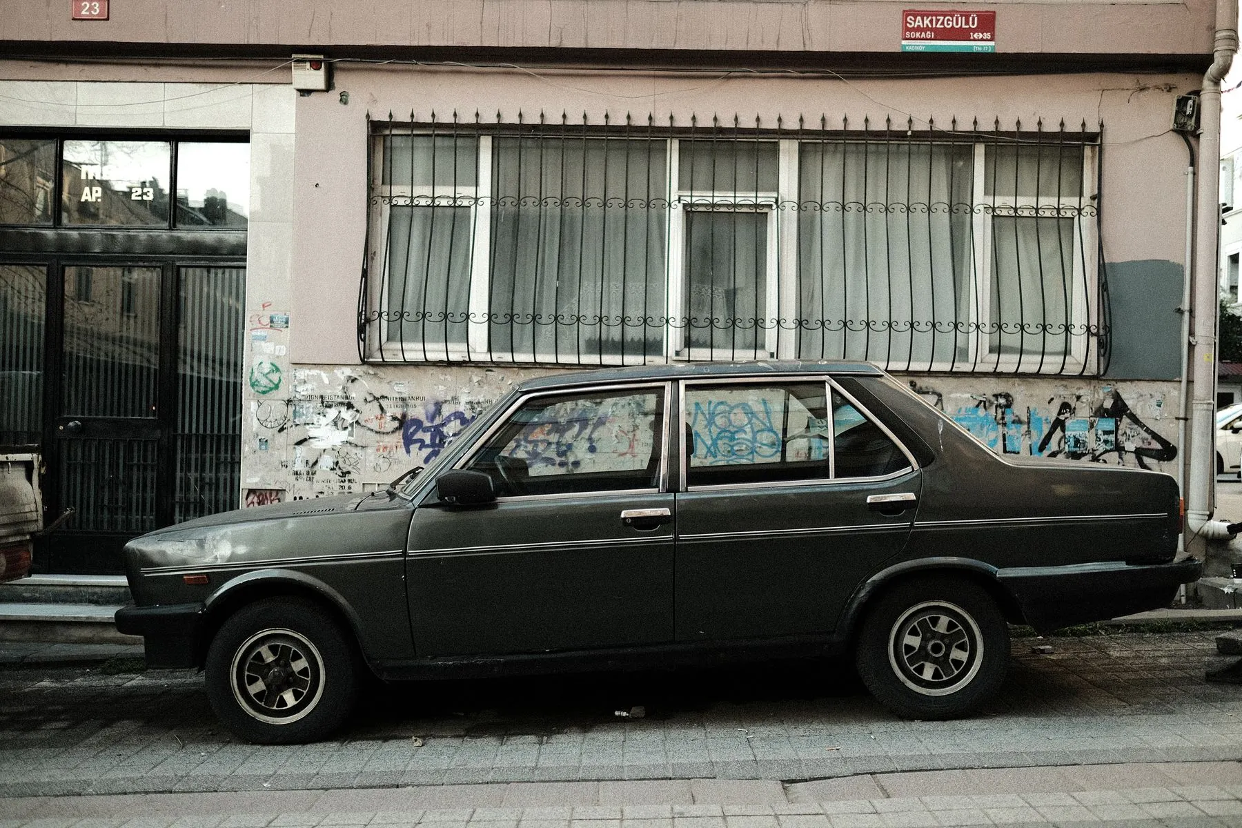 An old dark sedan parked at the curb in front of a building with barred windows