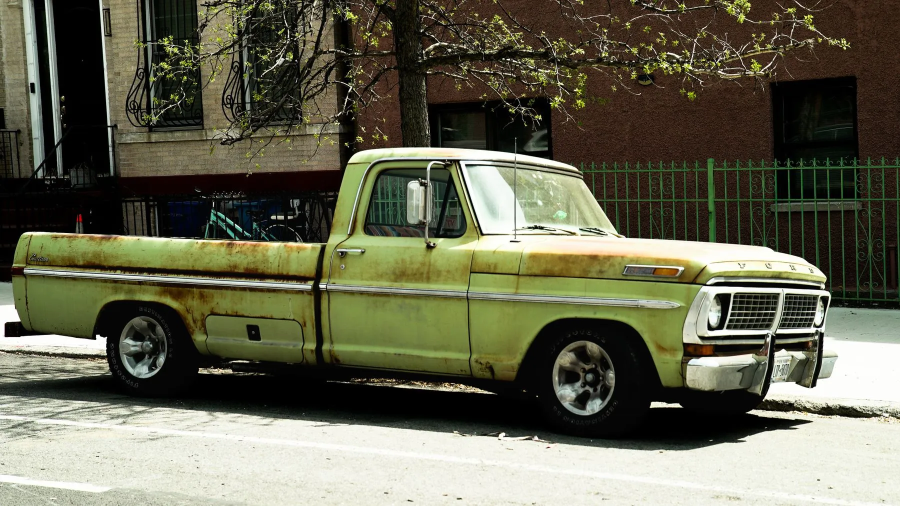 A faded green vintage pickup truck with rust on the fenders parked on a city street