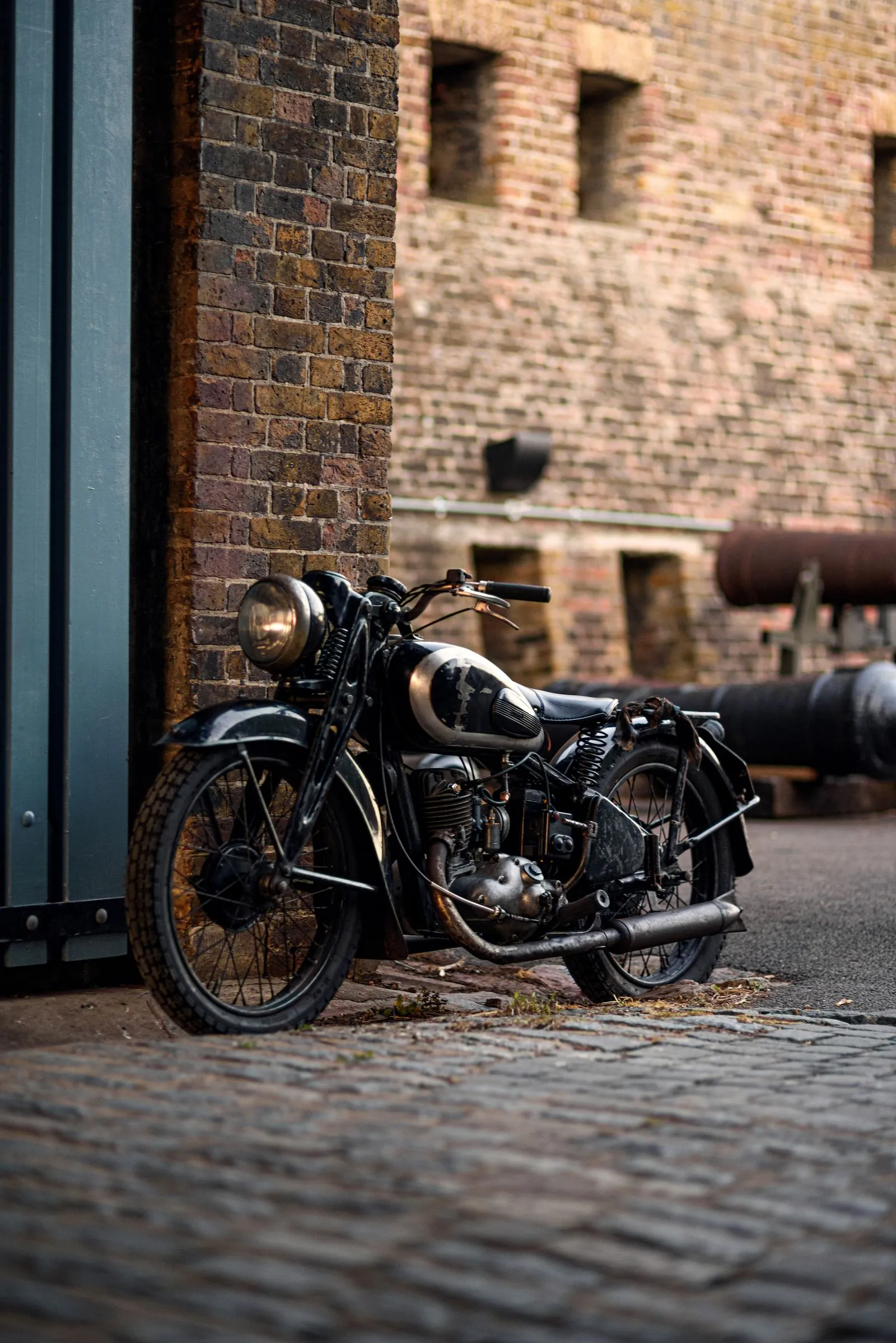 An old black motorcycle with a round headlight parked on cobblestones beside a brick building